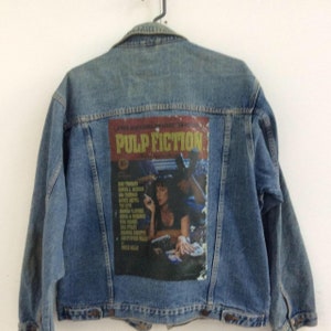 May include: A blue denim jacket with a Pulp Fiction movie poster patch on the back. The poster features Uma Thurman and John Travolta.