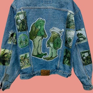 May include: A blue denim jacket with multiple embroidered patches featuring illustrations of frogs in various scenes. The patches are sewn onto the back of the jacket.