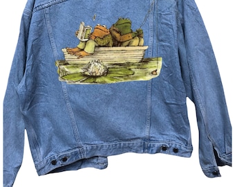 May include: A light blue denim jacket with a printed image of two frogs in a boat on the back. The frogs are wearing clothes and one is reading a book.
