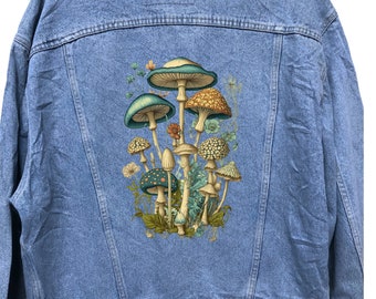 May include: Light blue denim jacket featuring a colorful mushroom graphic on the back. The illustration includes various mushroom types in shades of green, brown, and beige, set against a backdrop of foliage. The jacket has a classic button-up design.
