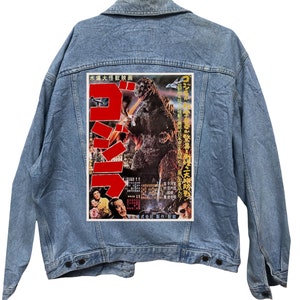 Può includere: Una giacca di jeans blu con una toppa di un poster di un film vintage di Godzilla sul retro. Il poster presenta un grande carattere giapponese rosso per "Godzilla" e un'immagine in bianco e nero del mostro.