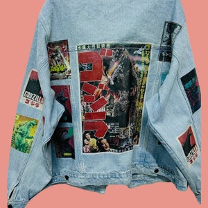 May include: A light blue denim jacket with a variety of Godzilla movie posters printed on the back. The posters are in Japanese and feature images of the monster Godzilla.