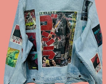 May include: A light blue denim jacket with a variety of Godzilla movie posters printed on the back. The posters are in Japanese and feature images of the monster Godzilla.