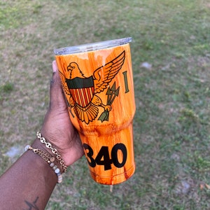 May include: A brown and orange wood-grain tumbler with a clear lid. The tumbler features a graphic of the Virgin Islands flag with an eagle and the number 340.