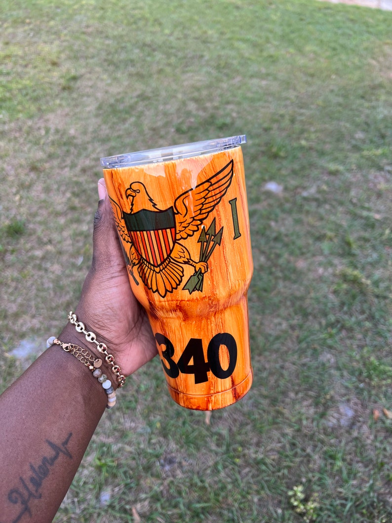 May include: A brown and orange wood-grain tumbler with a clear lid. The tumbler features a graphic of the Virgin Islands flag with an eagle and the number 340.