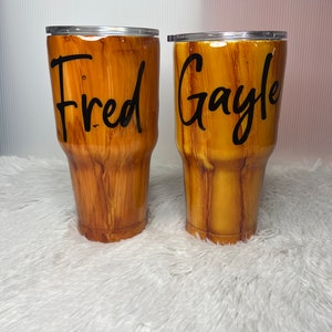 May include: Two brown and gold wood-grain tumblers with lids. The tumbler on the left has the name "Fred" written on it. The tumbler on the right has the name "Gayle" written on it.