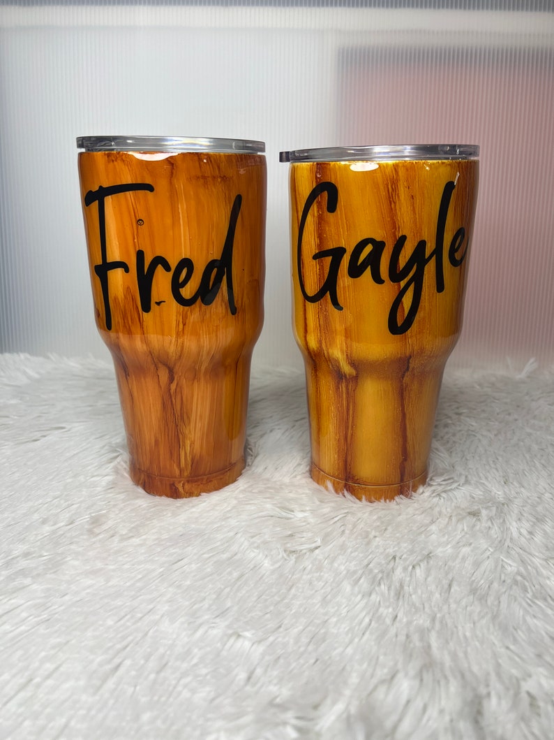 May include: Two brown and gold wood-grain tumblers with lids. The tumbler on the left has the name "Fred" written on it. The tumbler on the right has the name "Gayle" written on it.