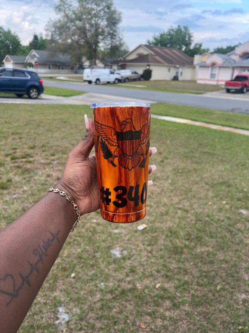 May include: A brown and gold stainless steel tumbler with a wood grain design. The tumbler features a black and white image of the coat of arms of the United States Virgin Islands and the number "#340" printed on the side.