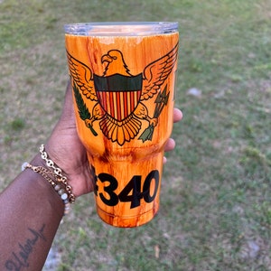May include: A brown and orange wood-grain tumbler with a gold eagle and shield design. The shield has a red, white, and blue striped pattern. The tumbler has a black number "340" on the bottom.
