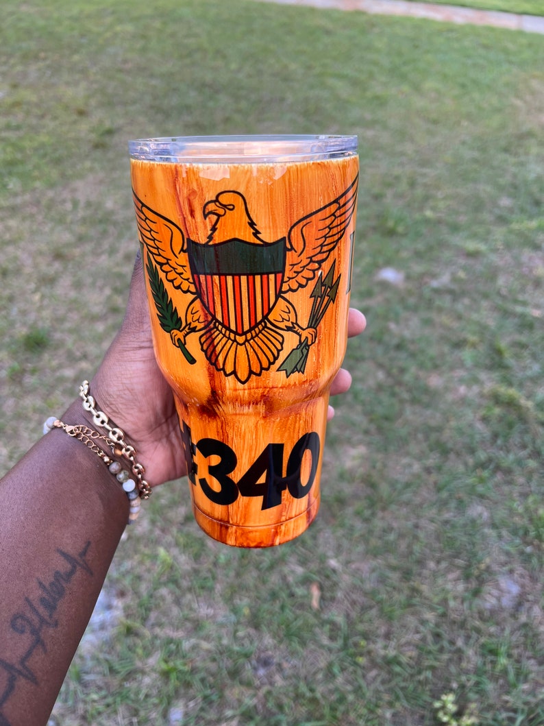May include: A brown and orange wood-grain tumbler with a gold eagle and shield design. The shield has a red, white, and blue striped pattern. The tumbler has a black number "340" on the bottom.