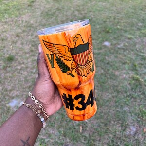 May include: A brown and orange stainless steel tumbler with a wood grain design. The tumbler features a green and red eagle with a shield and the number 34 on the side.