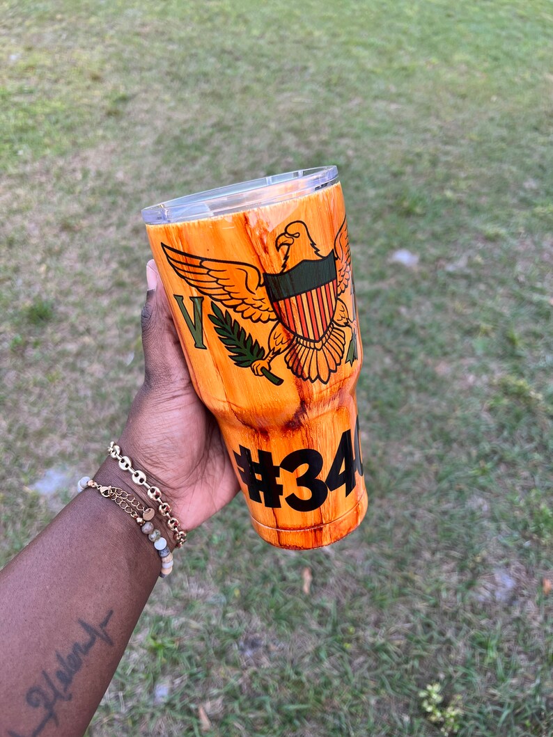 May include: A brown and orange stainless steel tumbler with a wood grain design. The tumbler features a green and red eagle with a shield and the number 34 on the side.