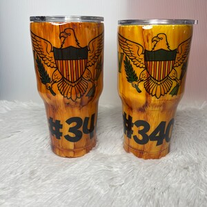 May include: Two brown and gold stainless steel tumblers with a wood grain design. The tumblers feature the flag of the US Virgin Islands and the numbers #34 and #340.