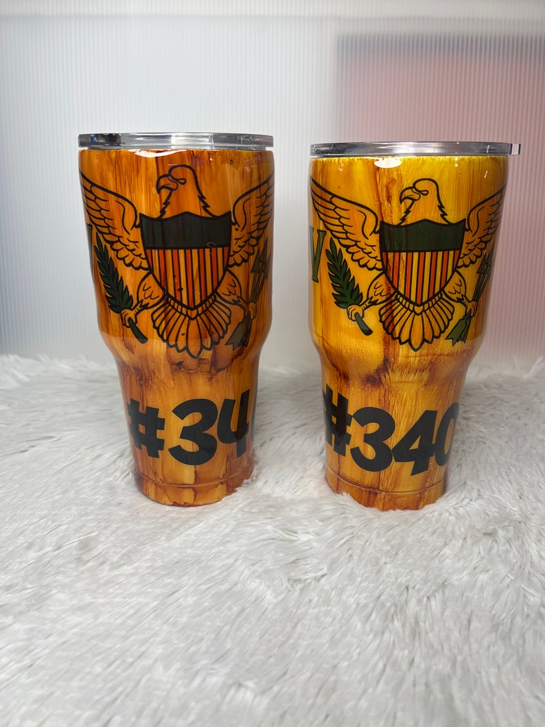 May include: Two brown and gold stainless steel tumblers with a wood grain design. The tumblers feature the flag of the US Virgin Islands and the numbers #34 and #340.