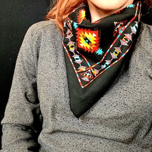 May include: A black square scarf with a colorful geometric pattern in red, yellow, turquoise, and white. The scarf is worn around the neck, with the pattern prominently displayed. The scarf is made of a soft material.