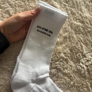 Könnte beinhalten: Weiße gerippte Socken mit dem Text "ICH ATME EIN. ICH RASTE AUS." in Schwarz. Die Socken sind aus einem weichen Material und sind umgeschlagen.