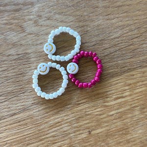 Könnte beinhalten: Drei Perlenringe mit Smiley-Designs. Zwei Ringe sind weiß mit weißen Perlen und gelben Smileys. Der dritte Ring ist magenta mit einem gelben Smiley.