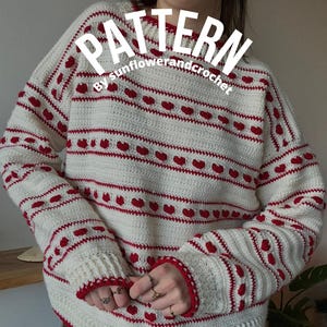 Peut inclure: Pull en crochet crème avec un motif de cœurs et de rayures rouges. Le pull à manches longues a des poignets et un ourlet côtelés. Le mot "PATTERN" est affiché en blanc en haut, avec "by sunflowerandcrochet" en dessous.