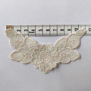 Op de afbeelding: Een witte kant applique met een bloemenmotief, ongeveer 18 cm breed. De applique heeft een grote centrale bloem met kleinere bloemen en bladeren eromheen.