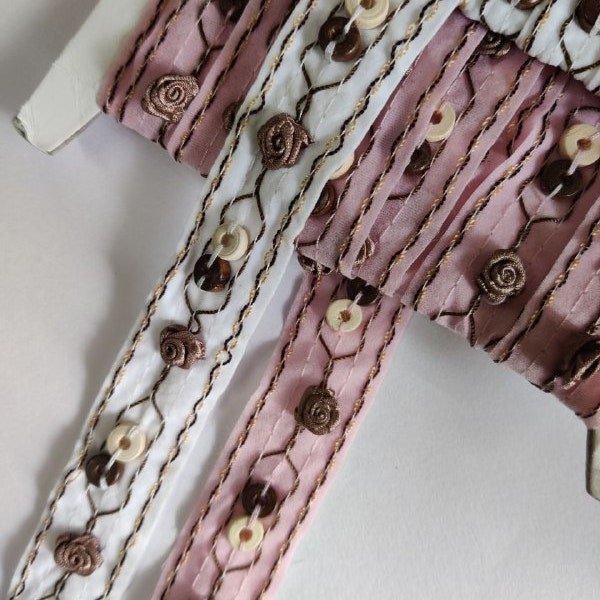 Op de afbeelding: Twee spoelen met witte en roze bies met bruine en beige knopen en geborduurde bloemen. De bies is gemaakt van een transparante stof en heeft een delicate, vrouwelijke uitstraling.