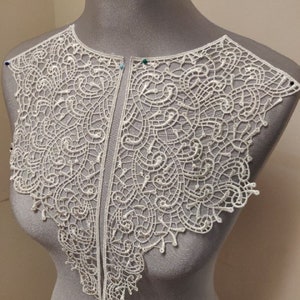 May include: White lace collar with a delicate floral pattern. The collar is made of two separate pieces that are joined together at the center.