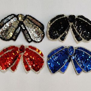 May include: Four sequined bow appliques in different colors: silver, black, red, and blue. Each bow is made of small, round sequins sewn together in a bow shape.