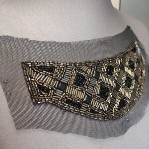 May include: A black and silver beaded embellishment on a sheer black fabric. The embellishment is in the shape of a curved, abstract design.