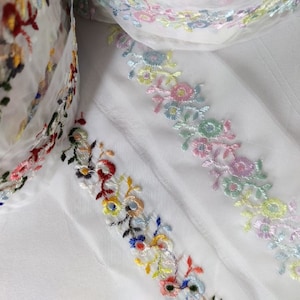 May include: White sheer fabric with two rows of colourful embroidered floral trim. The trim features a variety of colours including red, blue, green, yellow, and pink.