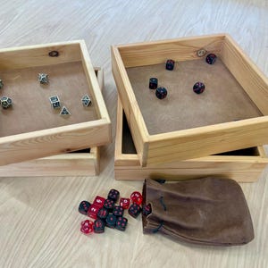 May include: Two square wooden dice trays with light brown wood grain frames and tan felt bases. One tray contains five multi-sided dice, the other contains five six-sided dice. A brown leather drawstring pouch sits nearby with loose red and black dice.