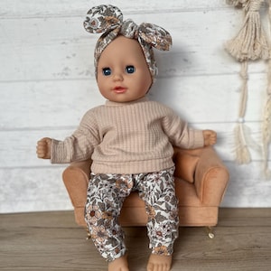 May include: A doll wearing a floral headband, a light pink sweater, and floral pants. The doll is sitting on a small, light brown velvet armchair.