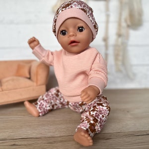 May include: A doll wearing a pink and brown floral patterned outfit with a pink beanie. The doll is sitting on a wooden surface.