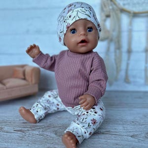 Eine Puppe in einem mauvefarbenen, gerippten Langarmshirt, einer Hose mit Blumenmuster und einer passenden Mütze.  Das Outfit hat einen weißen Hintergrund mit mauve und grünen Blumenmustern.