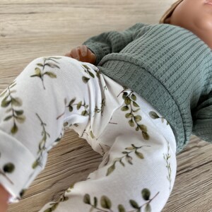 May include: White baby leggings with a green leafy print. The leggings are paired with a green ribbed sweater.