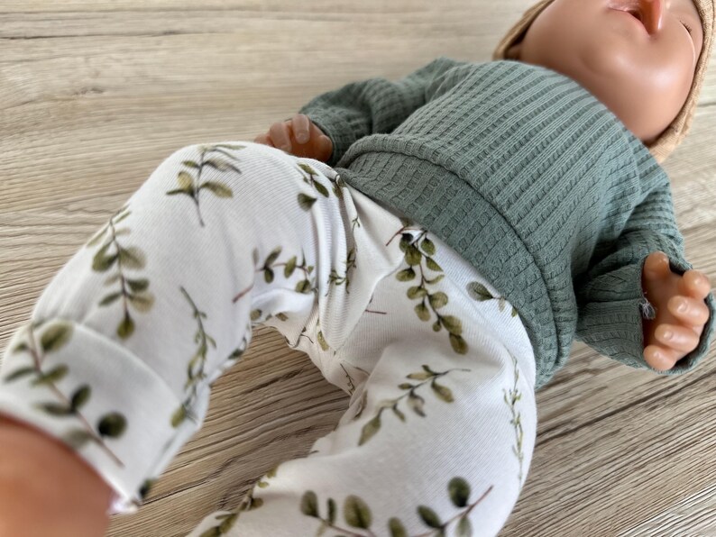 May include: White baby leggings with a green leafy print. The leggings are paired with a green ribbed sweater.