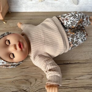 May include: A doll wearing a beige waffle knit long-sleeve shirt and floral print pants. The doll is wearing a floral print headband and has closed eyes.