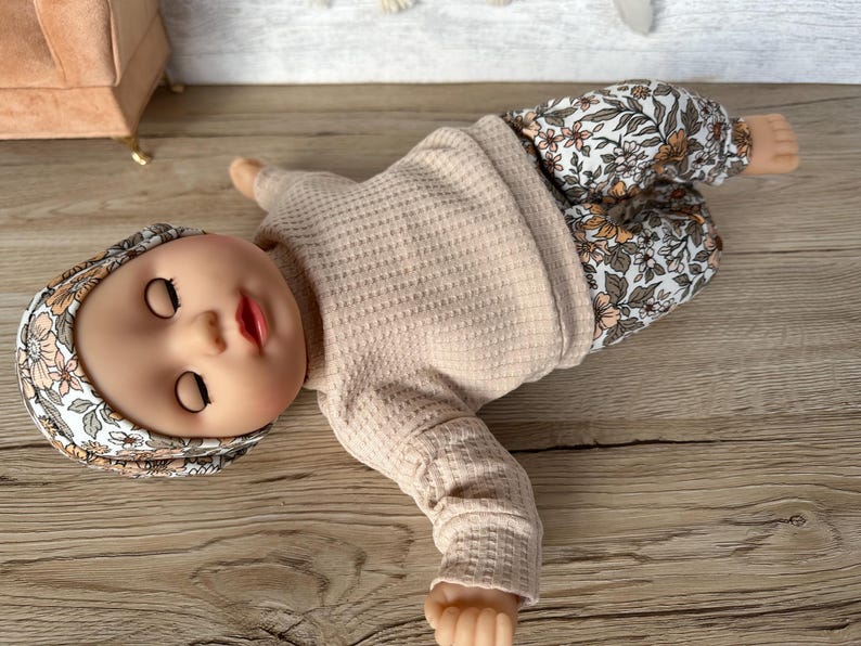 May include: A doll wearing a beige waffle knit long-sleeve shirt and floral print pants. The doll is wearing a floral print headband and has closed eyes.