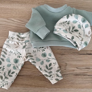 May include: A sage green ribbed long-sleeve top paired with matching trousers and a beanie, all featuring a white base with a green and brown botanical leaf pattern. The set includes a crew neck top, elastic waistband trousers, and a soft fabric beanie.