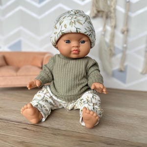 Pu&ograve; includere: Una bambola neonato indossa una maglia a maniche lunghe a coste verde oliva, pantaloni a coste abbinati con un motivo a foglie bianche e verdi e un berretto a coste coordinato con la stessa stampa. La bambola ha un incarnato liscio e occhi scuri.