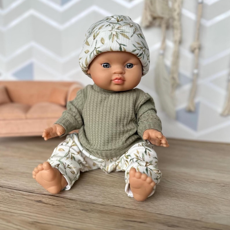 Pu&ograve; includere: Una bambola neonato indossa una maglia a maniche lunghe a coste verde oliva, pantaloni a coste abbinati con un motivo a foglie bianche e verdi e un berretto a coste coordinato con la stessa stampa. La bambola ha un incarnato liscio e occhi scuri.