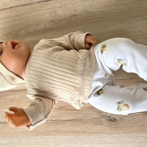Puede incluir: Una mu&ntilde;eca beb&eacute; con una camisa de manga larga de color beige con textura de canal&eacute; y pantalones blancos con estampado de lim&oacute;n.
