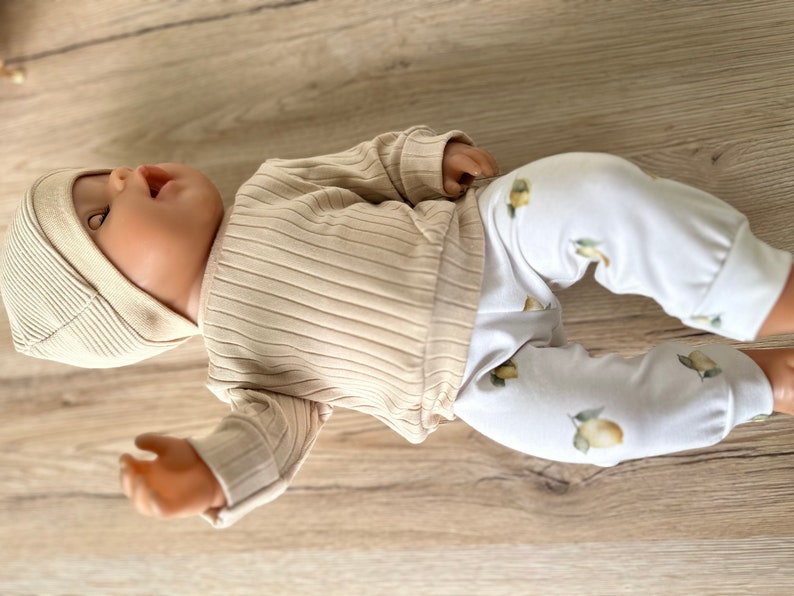 Puede incluir: Una mu&ntilde;eca beb&eacute; con una camisa de manga larga de color beige con textura de canal&eacute; y pantalones blancos con estampado de lim&oacute;n.