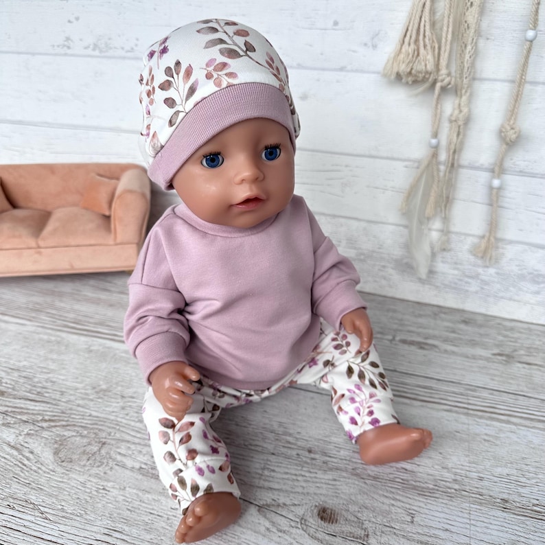 Eine Puppe trägt ein langärmeliges malvenfarbenes Sweatshirt, eine passende Hose mit weiß-braunem Blattmuster und eine dazu passende Mütze