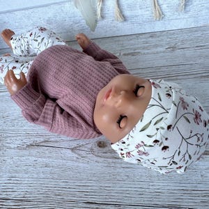May include: A doll wearing a mauve long-sleeved waffle-knit shirt, white pants with a brown and green floral pattern, and a matching white beanie with the same floral print. The doll has closed eyes and is positioned lying on its side.