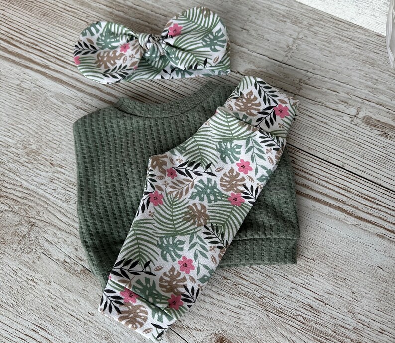 May include: A green waffle knit top and leggings set with a matching green and white floral headband. The leggings feature a white background with green leaves and pink flowers.