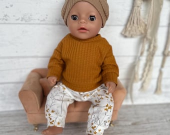 May include: A doll wearing a mustard yellow sweater and floral print pants, paired with a brown beanie. The doll is seated on a small, pink upholstered chair. The pants feature a white background with a delicate pattern of brown and yellow flowers.