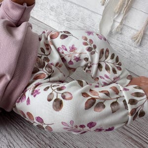 May include: A pair of white leggings featuring a botanical print of brown and purple leaves and small flowers, paired with a solid dusty pink long-sleeved top. The leggings have cuffed ankles and a fitted silhouette, while the top has a ribbed collar.