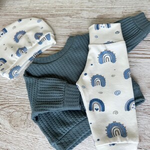 May include: A baby clothing set consisting of a solid dark teal long-sleeve shirt with a ribbed texture, paired with white pants and a matching white beanie, both featuring a repeating pattern of blue rainbows and small heart accents.