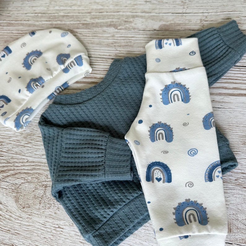 May include: A baby clothing set consisting of a solid dark teal long-sleeve shirt with a ribbed texture, paired with white pants and a matching white beanie, both featuring a repeating pattern of blue rainbows and small heart accents.