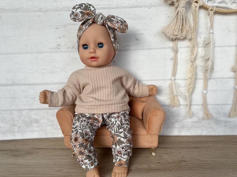 May include: A doll wearing a floral headband, a light pink sweater, and floral pants. The doll is sitting on a small, light brown velvet armchair.