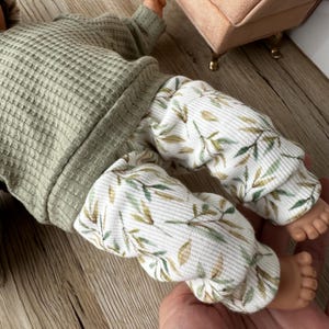 Pu&ograve; includere: Una bambola indossa una maglia a maniche lunghe in tessuto waffle color verde salvia e pantaloni a coste bianchi con un motivo di piccole foglie verdi e marroni. L'abbigliamento presenta polsini e orli, con la trama del tessuto ben visibile.